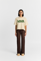 Woman wearing a beige t-shirt with green text and brown patterned pants on a white background